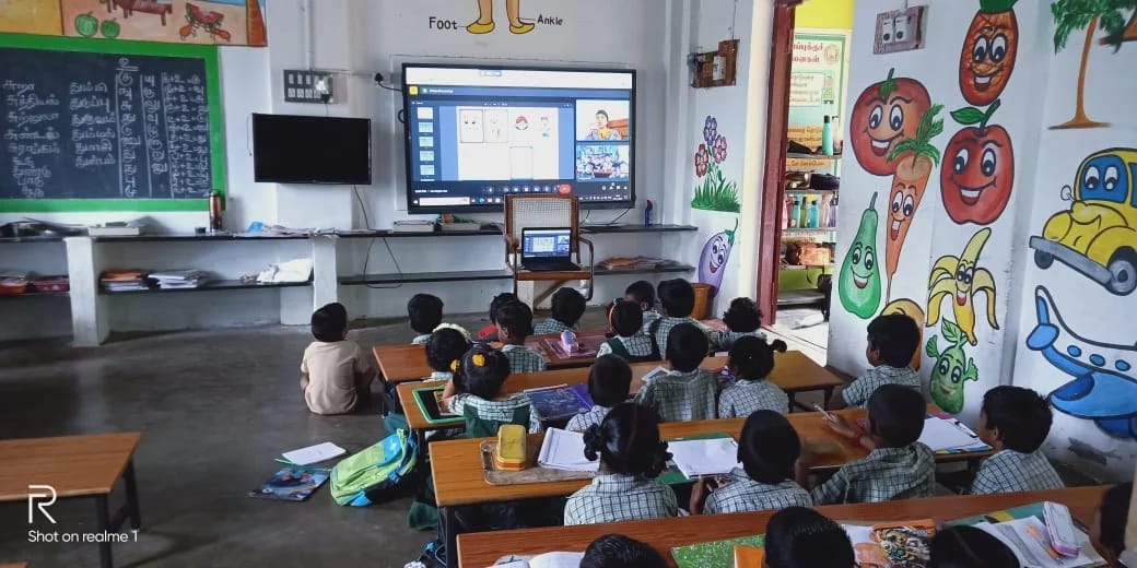 A class takes part in an online learning session displayed at the front of the room.