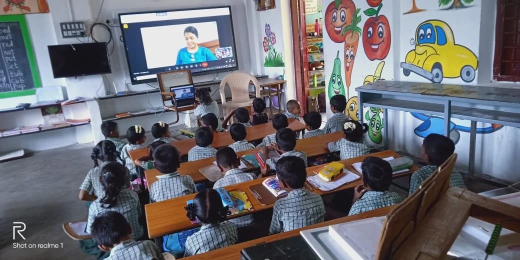 Children watch a reading session on a large classroom screen.
