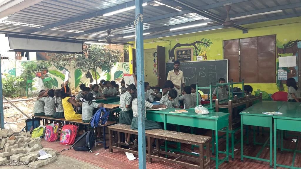 Children sit at green desks in an open school classroom.