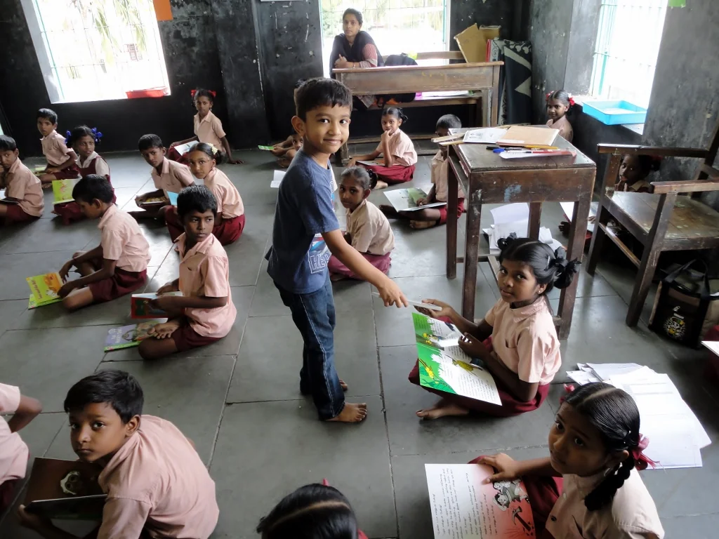 Vrishank holds a book while children gather around in a classroom.